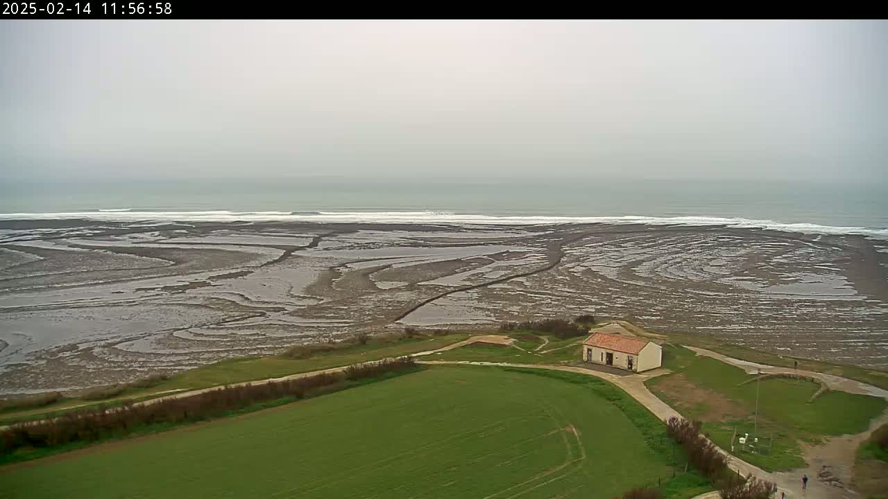 A small building sits on a grassy hill overlooking a wide expanse of exposed, patterned mudflats leading to a gray, overcast ocean with small waves.