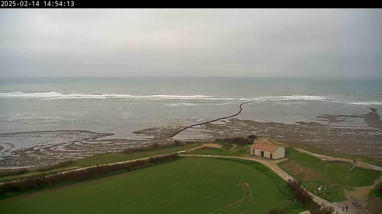 A small building sits on green land overlooking a grey, overcast ocean with waves breaking near a rocky outcropping.