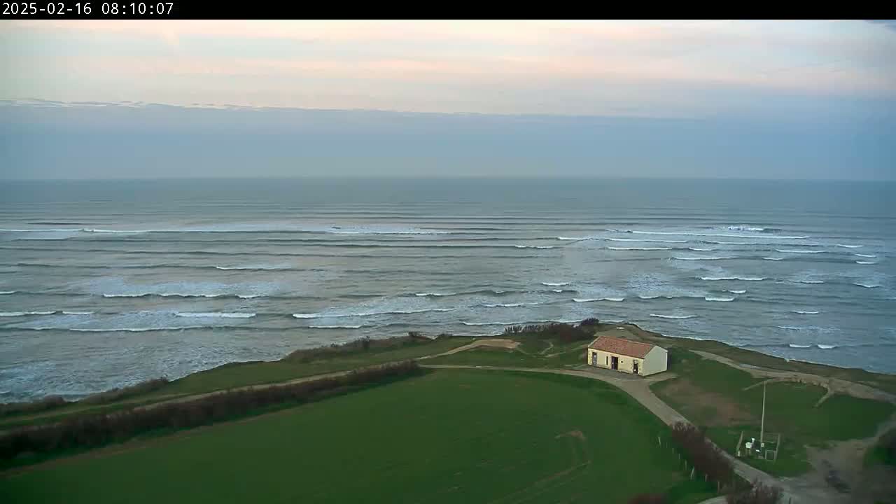 An aerial view shows a small building on a grassy cliff overlooking a calm ocean with rolling waves under a cloudy sky.