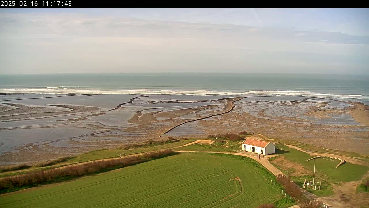 A small white building with a red roof sits on a grassy hill overlooking a tidal flat and a calm ocean with small waves under a partly cloudy sky.