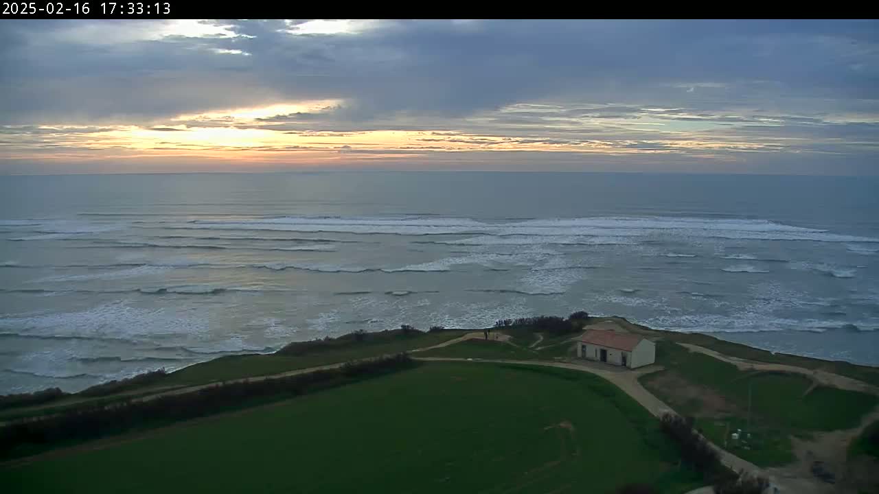 A small building sits on a grassy cliff overlooking a calm ocean with rolling waves under a mostly cloudy sunset sky.