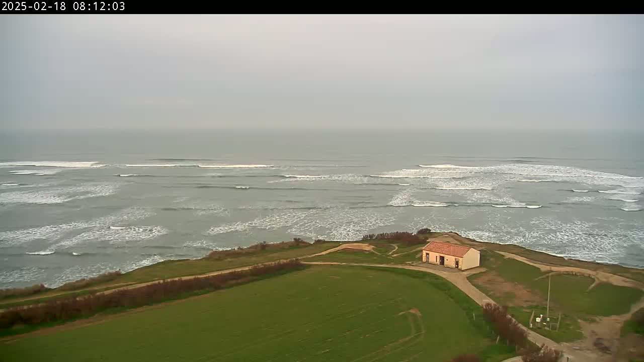 A small, tan building sits atop a grassy cliff overlooking a gray, overcast ocean with rolling waves.