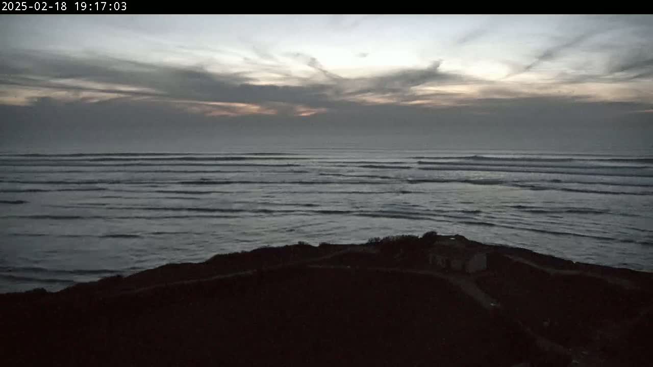 A dark, overcast sky hangs over a calm ocean with small waves rolling onto a silhouetted shoreline.