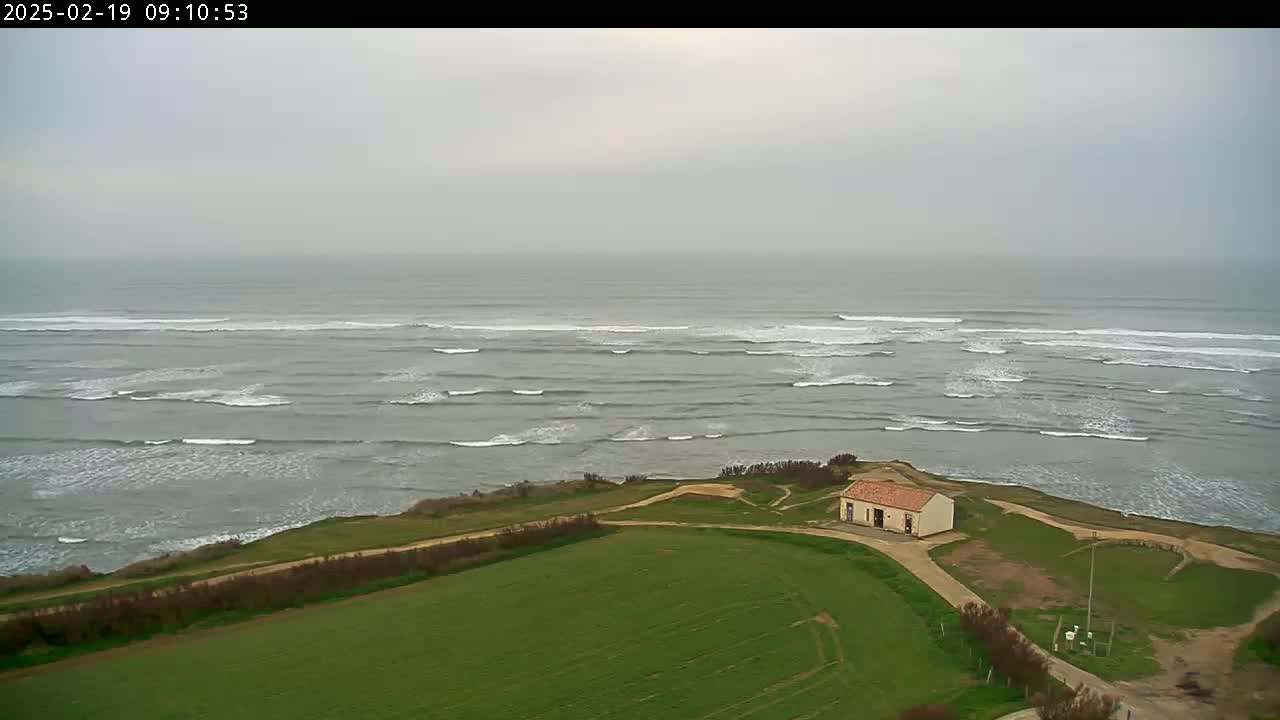A small building sits atop a grassy cliff overlooking a grey, overcast ocean with rolling waves.