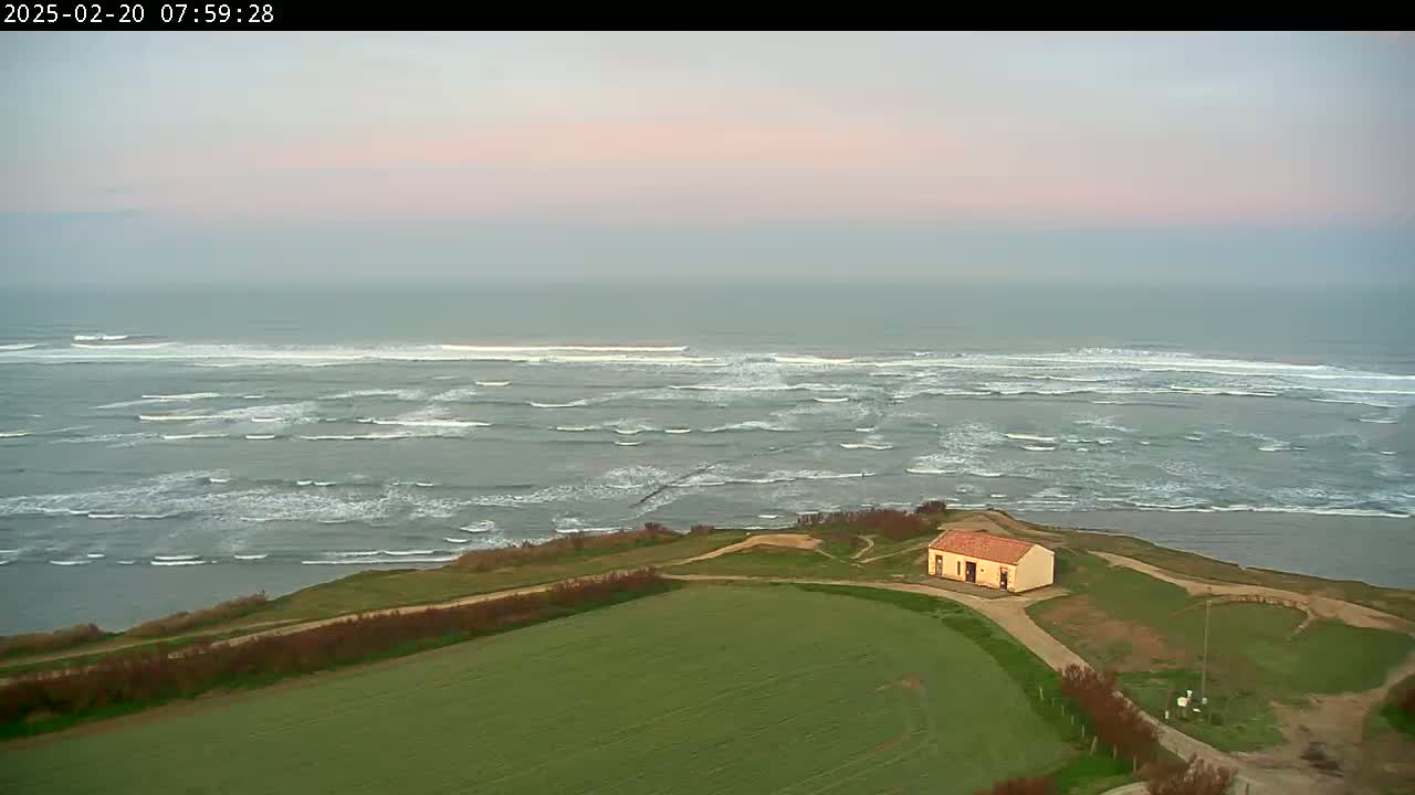 A small, tan building sits on a grassy cliff overlooking a gray ocean with moderate waves under a pale pink and gray sky.