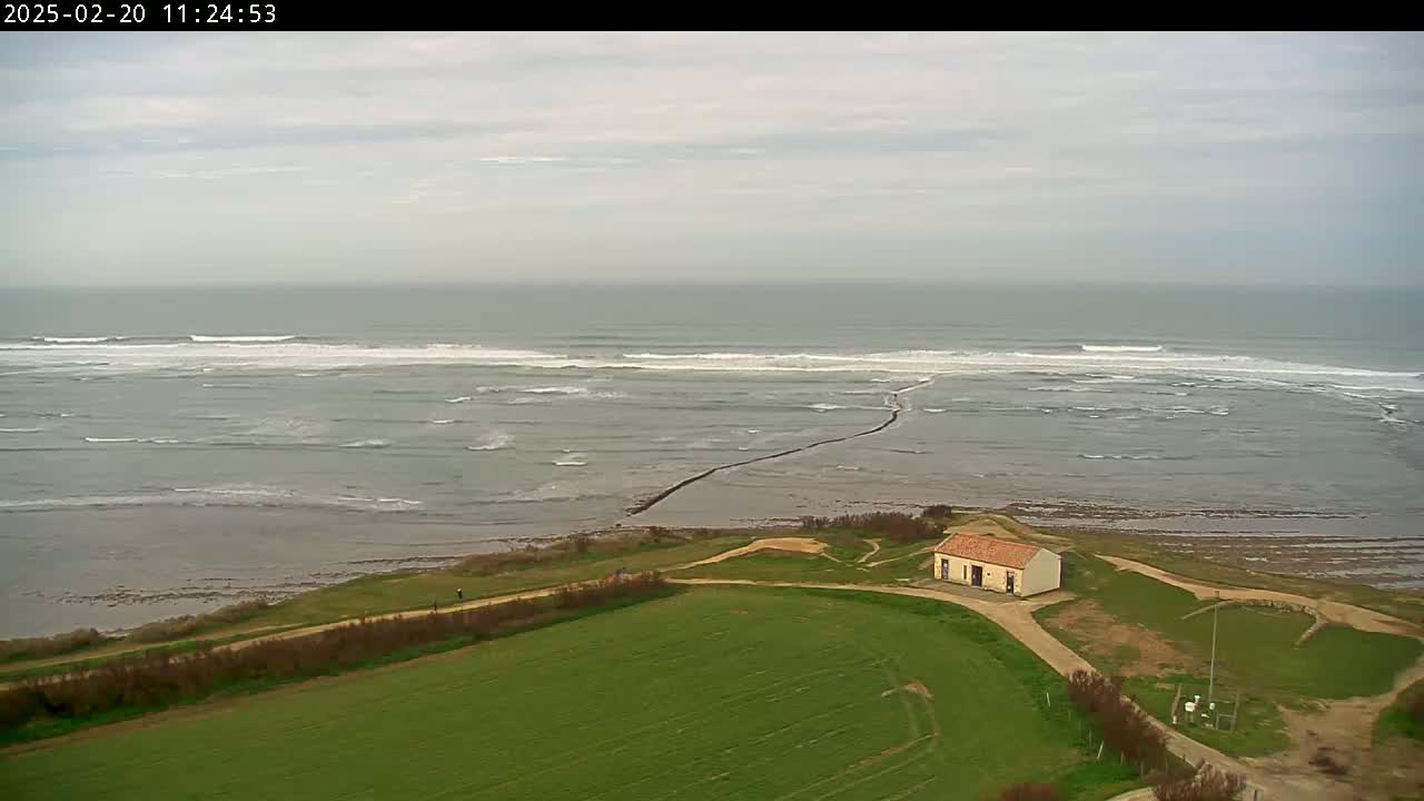 An aerial view shows a small building on a grassy coastal area overlooking a grey ocean with small waves under an overcast sky.