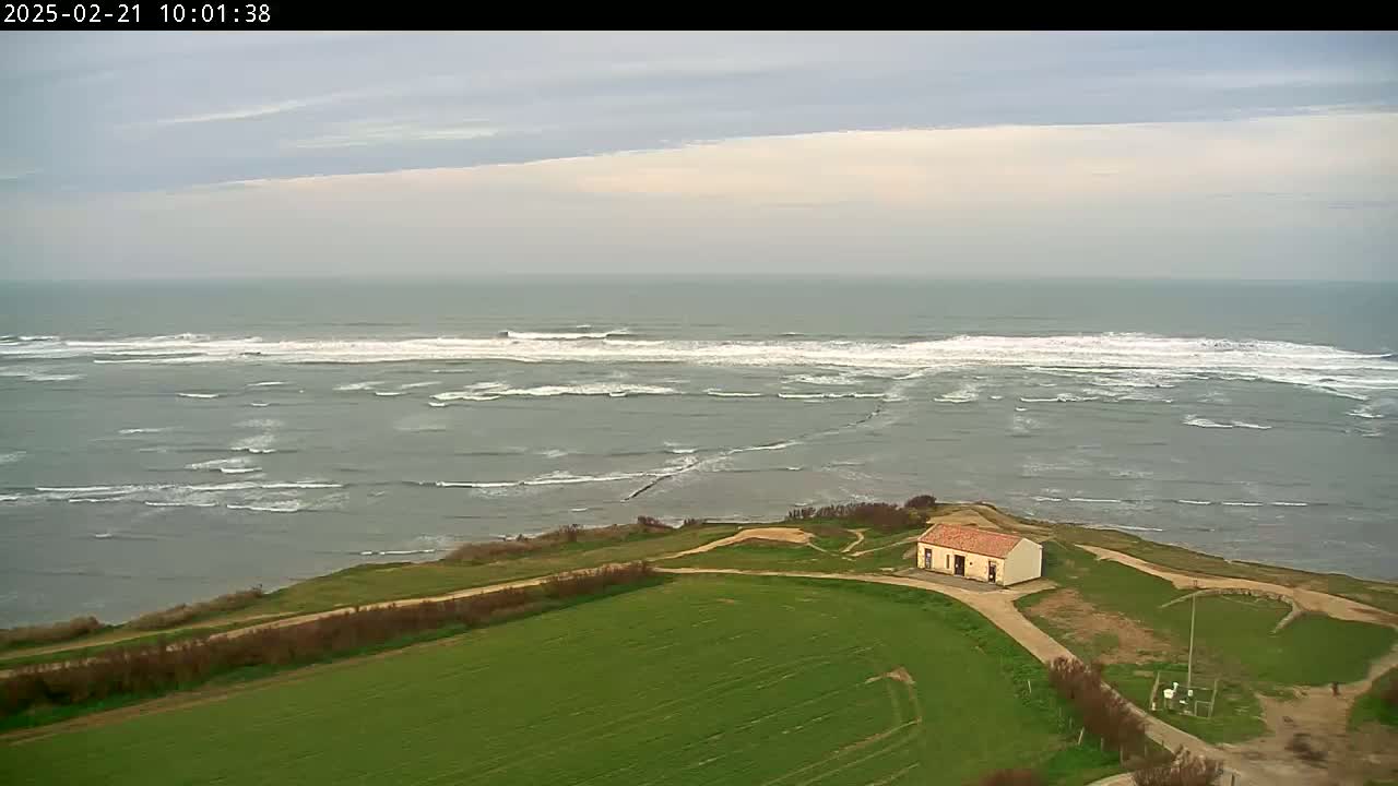 A small, light-colored building sits on a grassy cliff overlooking a gray, choppy sea with small waves under a mostly cloudy sky.