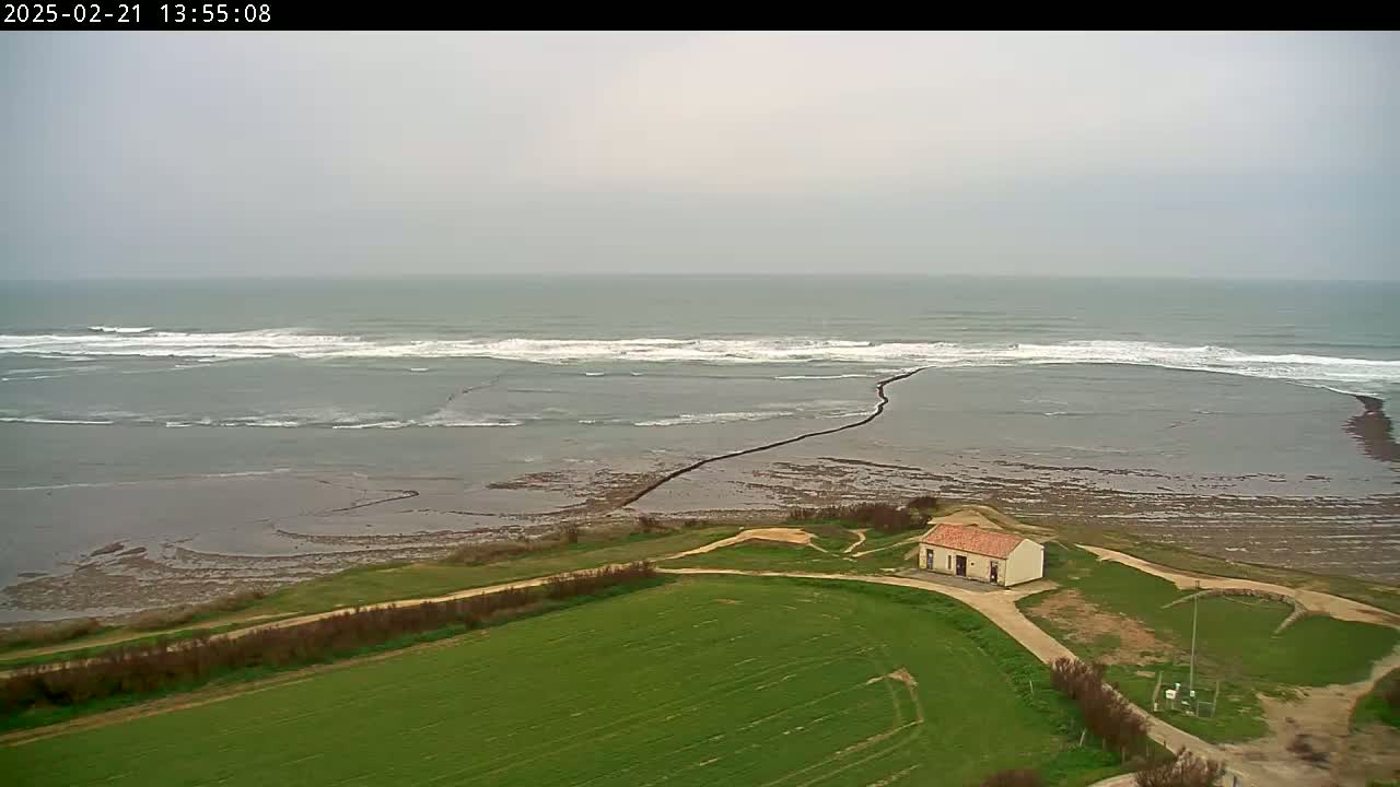 A small building sits on a grassy field overlooking a calm ocean with small waves under an overcast sky.
