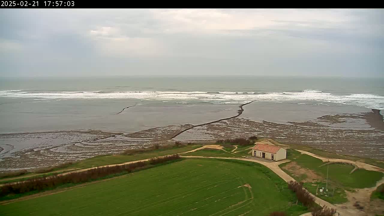 An aerial view shows a small building situated on a grassy area overlooking a coastline with exposed tidal flats and gentle waves under an overcast sky.