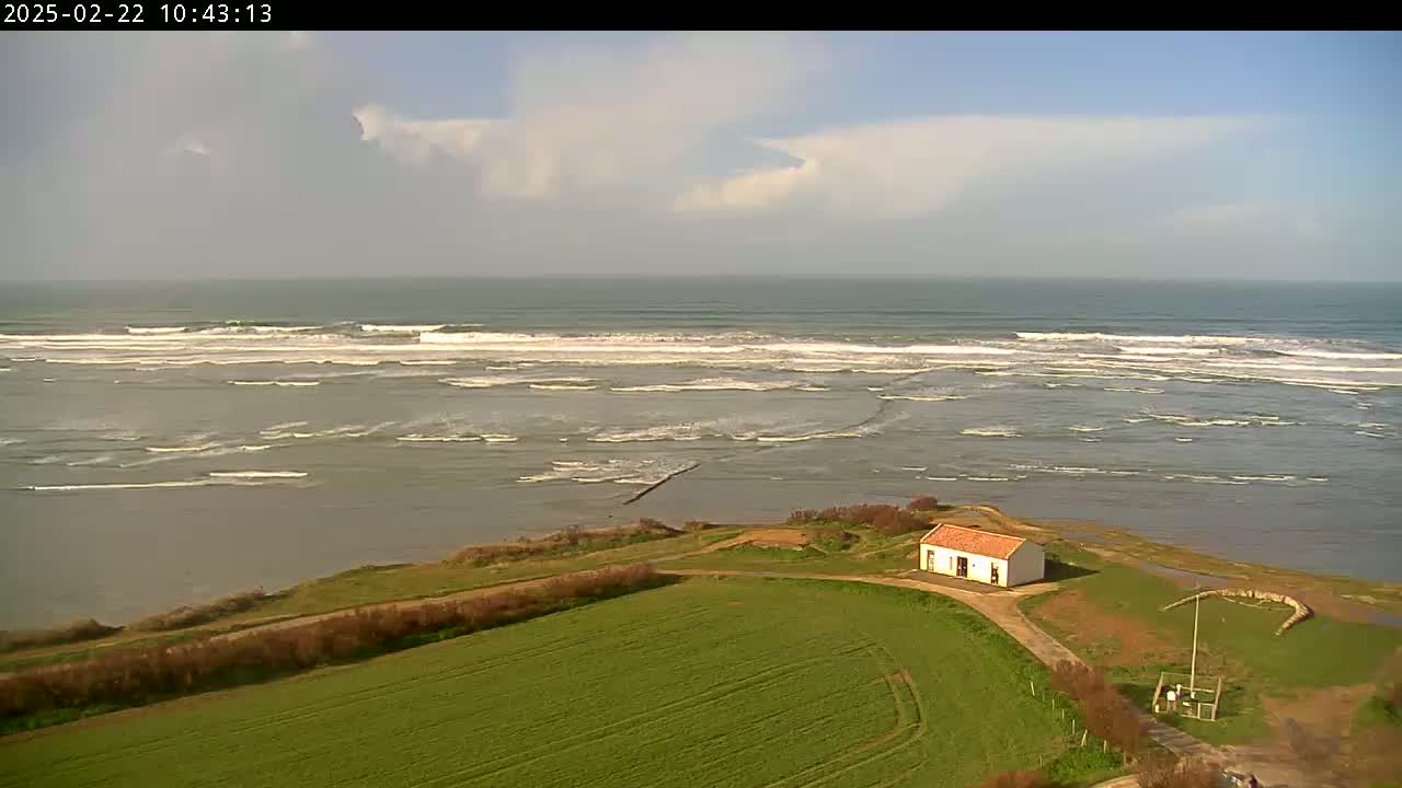 A small, white building with a red roof sits on a grassy field overlooking a calm bay with gentle waves breaking in the distance under a partly cloudy sky.