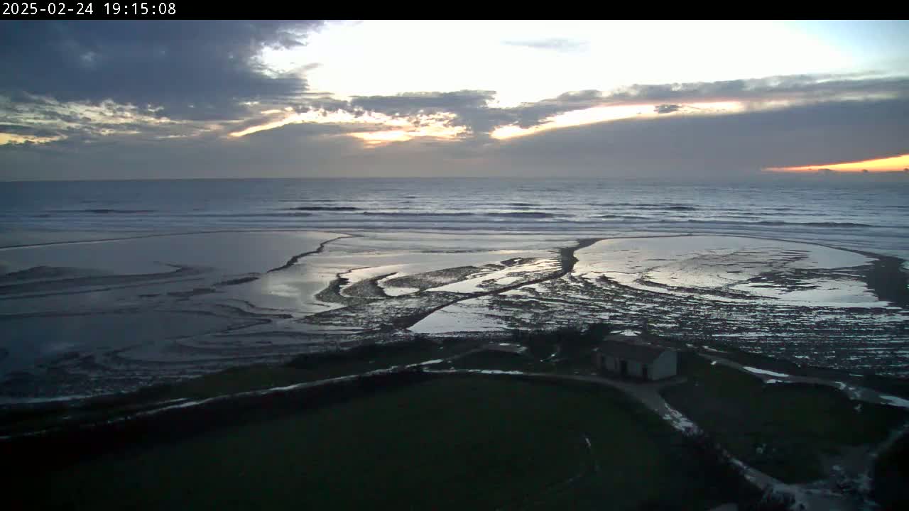 A coastal scene at sunset shows a calm ocean with small waves, a tidal flat with pools of water, and a small building on the grassy land above.