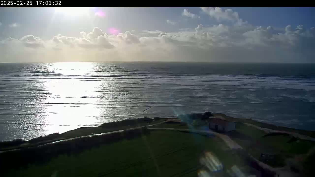 A partly cloudy sunset over a wavy ocean, viewed from a grassy clifftop with a small building visible below.
