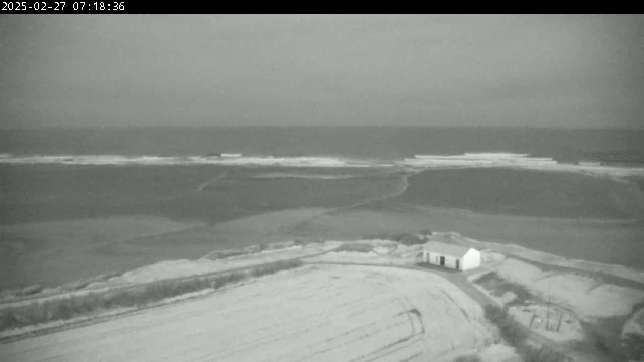 An overcast day shows a small, white building situated on a snow-covered field near a coastline with gentle waves.