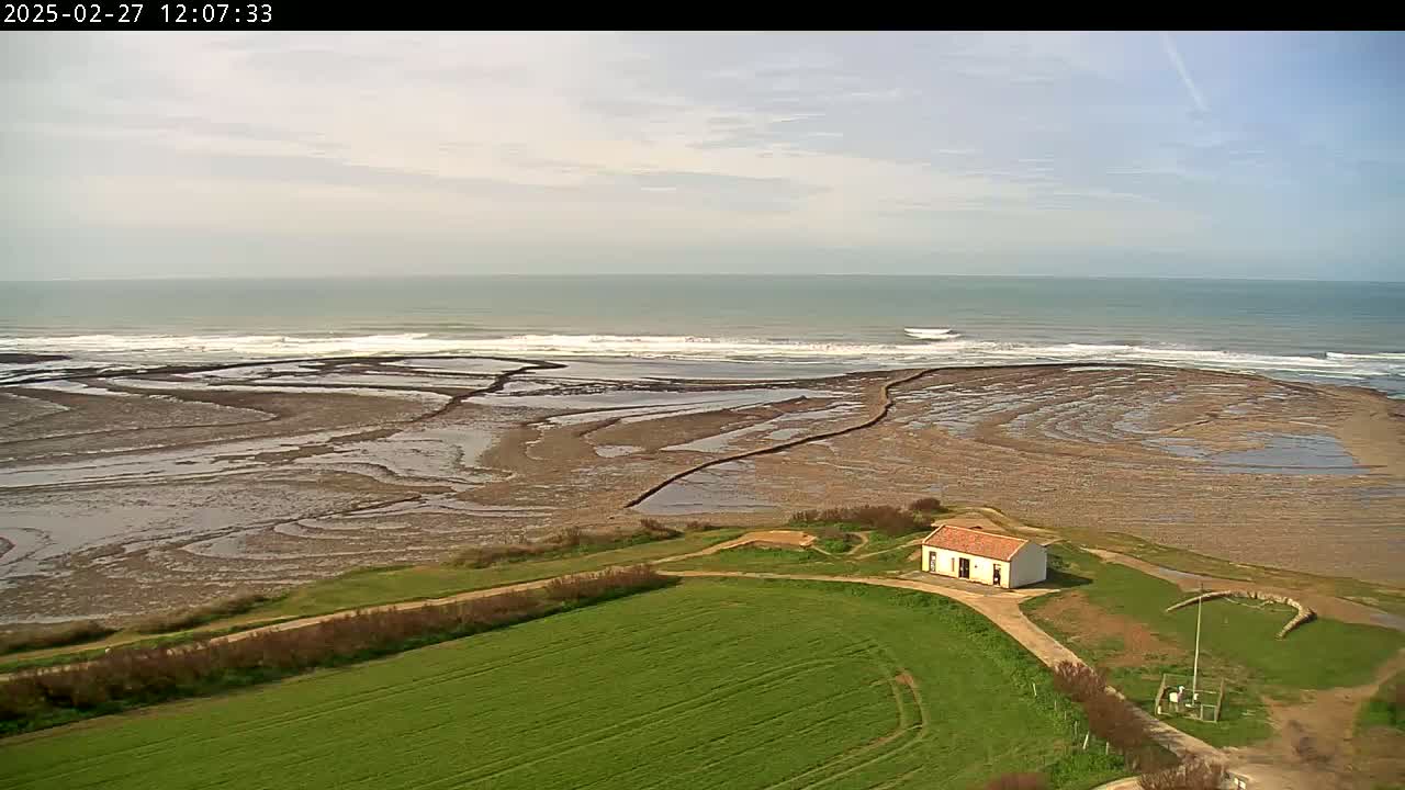 An aerial view shows a small, white building on a grassy area near a rocky, tidal shoreline with gentle waves under a partly cloudy sky.