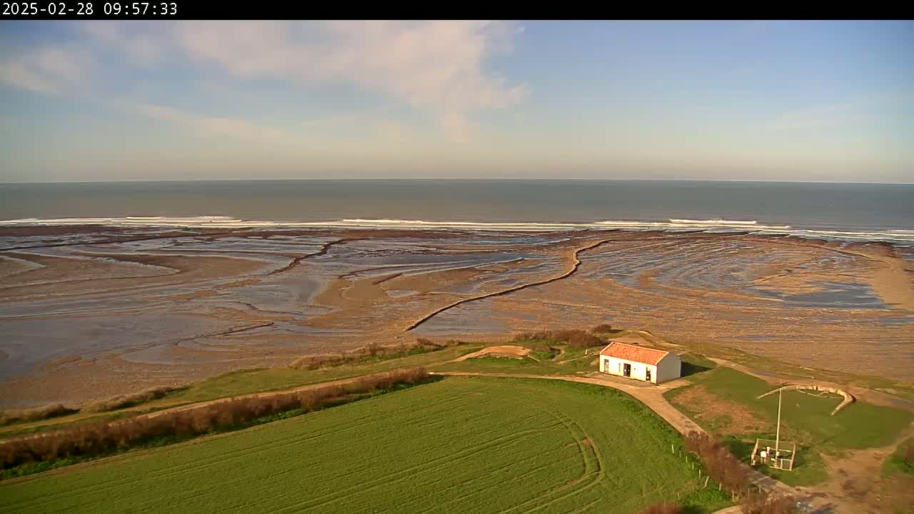 An aerial view shows a small building near a green field overlooking a tidal flat and ocean with small waves under a mostly clear sky.