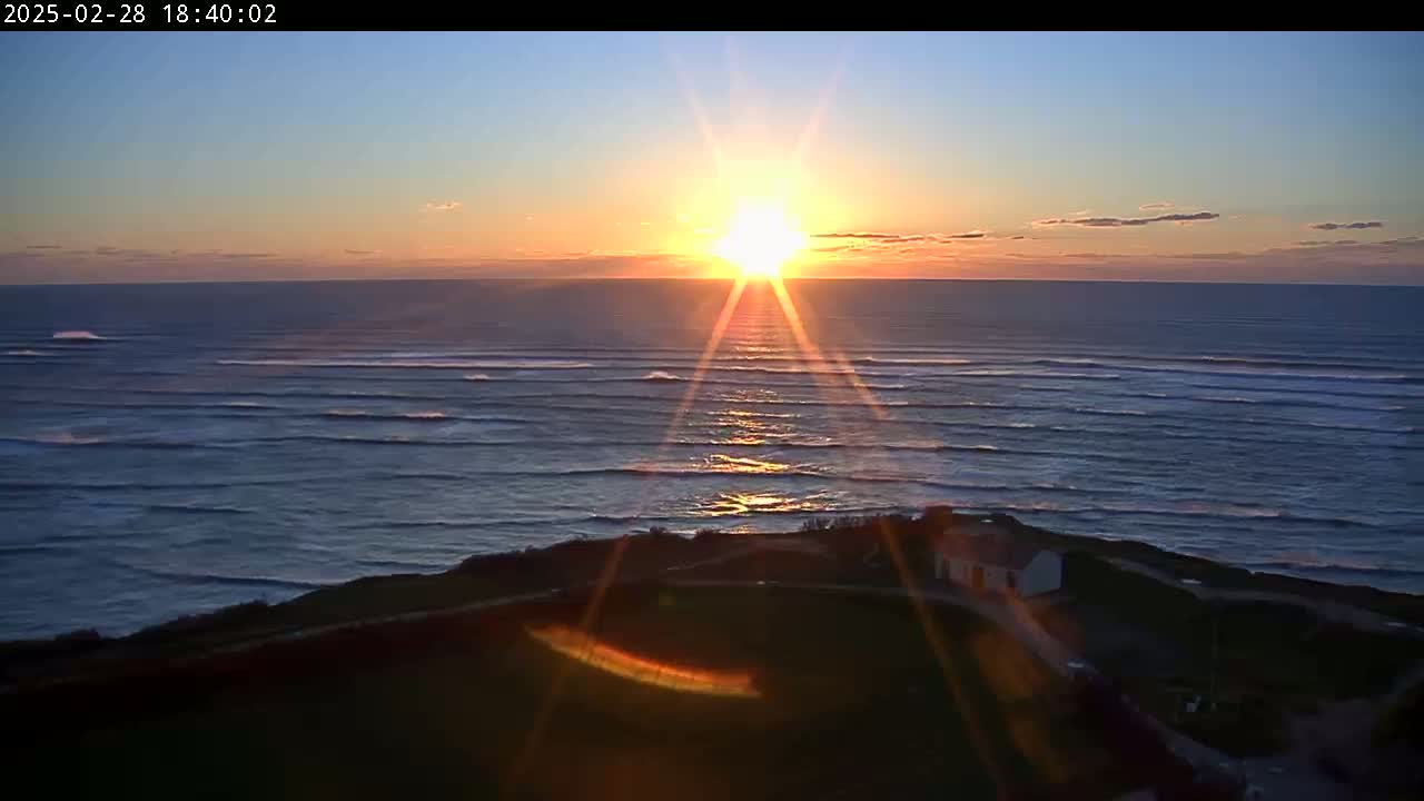 A sunset over a calm ocean with small waves rolling onto the shore, viewed from a grassy clifftop with a small building visible.