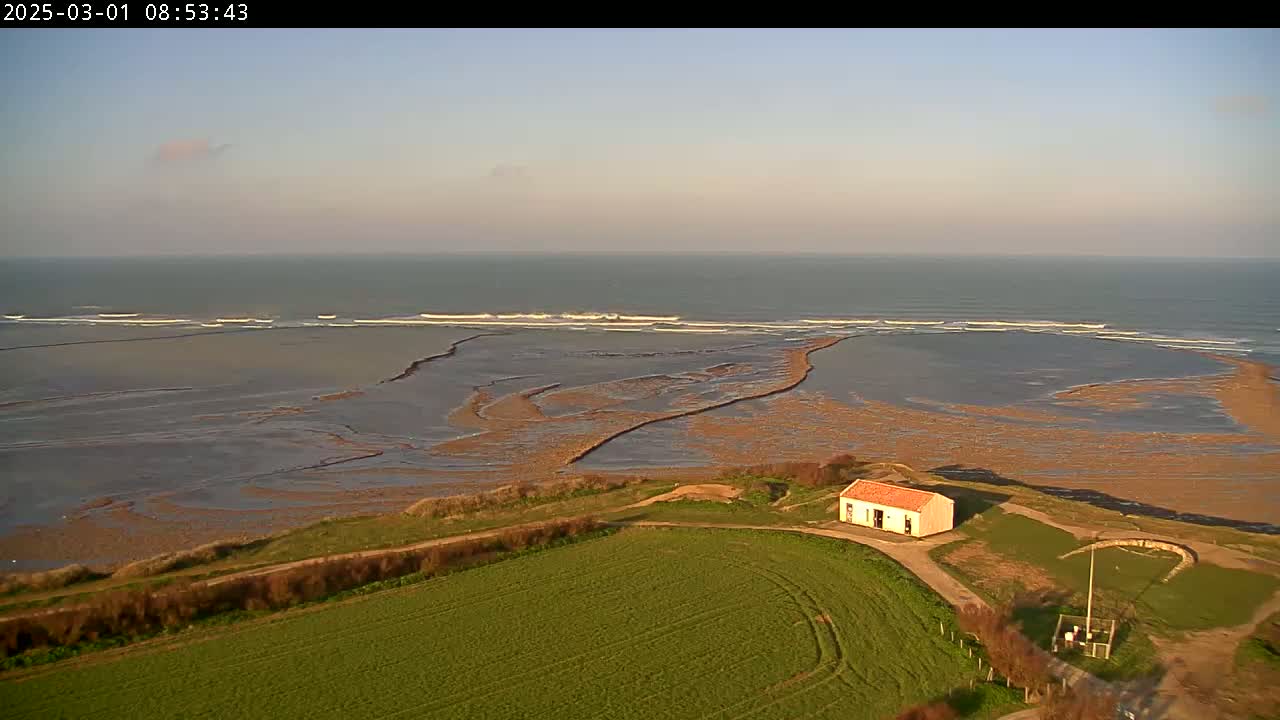 A small, single-story building sits atop a grassy hill overlooking a tidal flat and ocean with small waves under a mostly clear sky.
