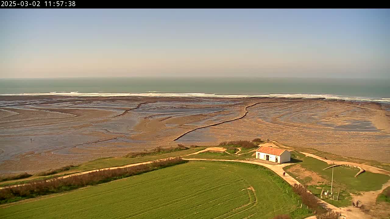 A small white building with a red roof sits on a grassy hill overlooking a vast, low-tide, patterned beach and a calm ocean under a clear sky.