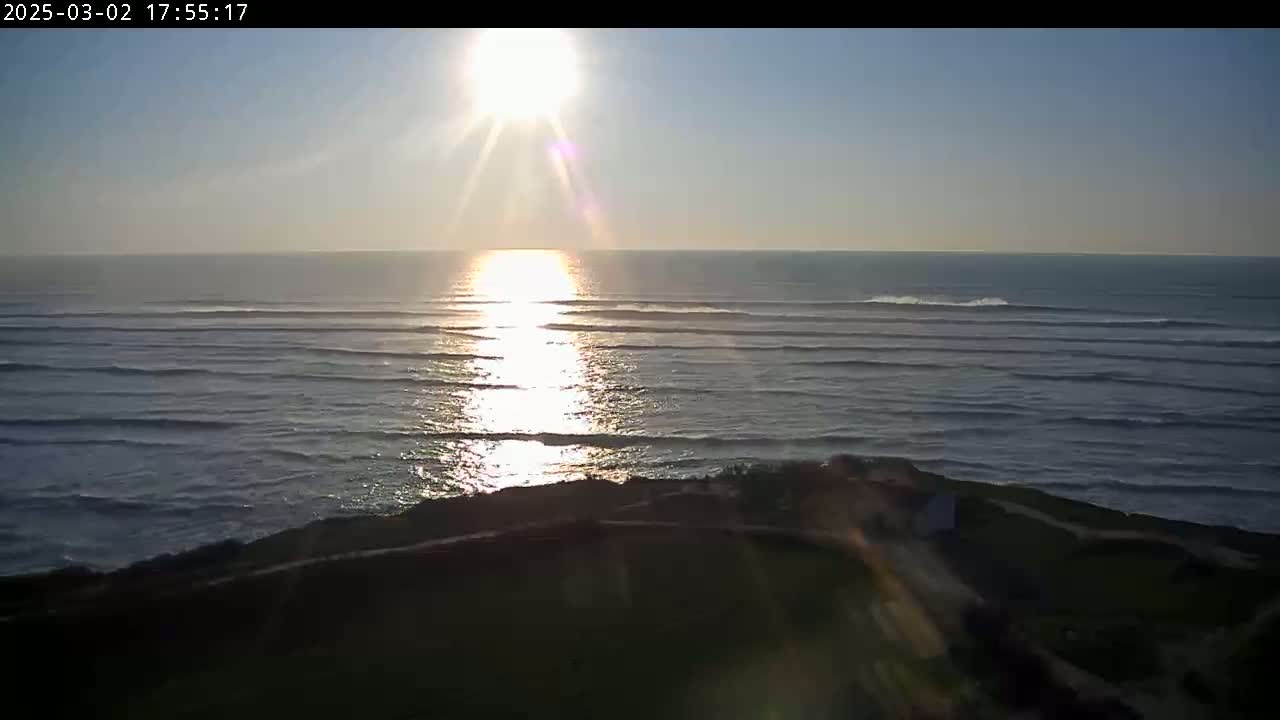 The setting sun shines brightly on a calm ocean with gently rolling waves, viewed from a dark, grassy hilltop.