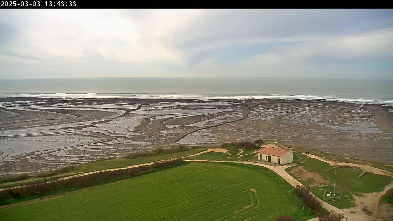 An aerial view shows a small building near a coastline under partly cloudy skies, with an expanse of tidal flats extending to a gently rolling ocean.