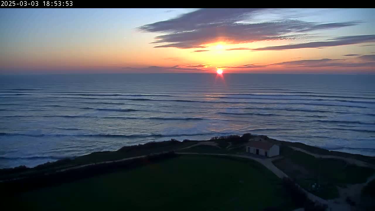 A sunset over a dark ocean with rolling waves and a small building on a grassy cliff.