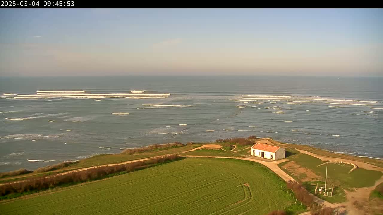 A small, single-story building sits atop a grassy hill overlooking a calm ocean with small waves breaking in the distance under a clear sky.