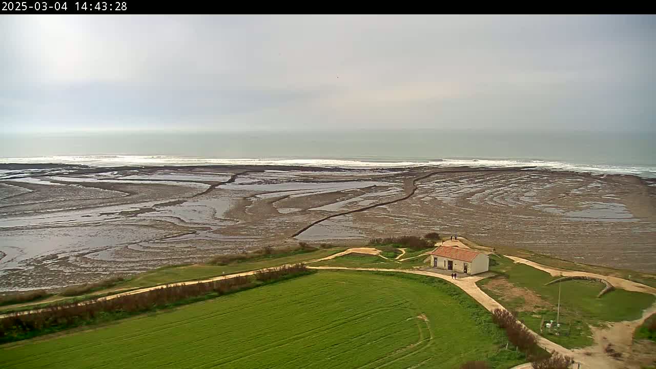 A small building sits on a grassy area overlooking a wide expanse of tidal flats leading to a grey, overcast ocean with small waves.