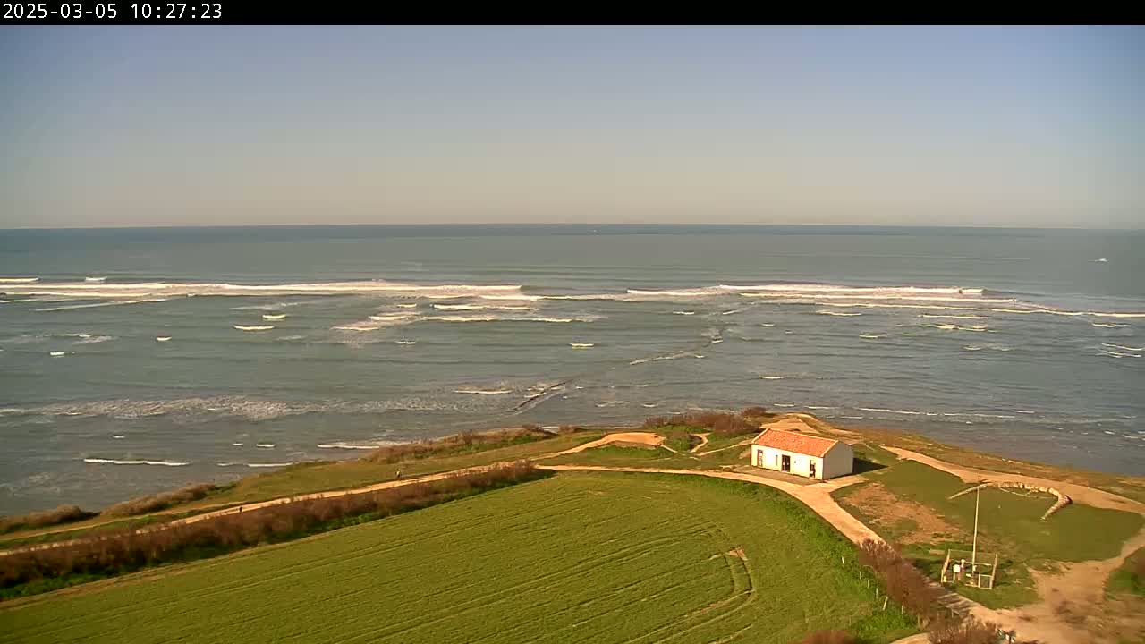 A small white building with a red roof sits on a grassy hill overlooking a calm ocean with small waves under a clear sky.