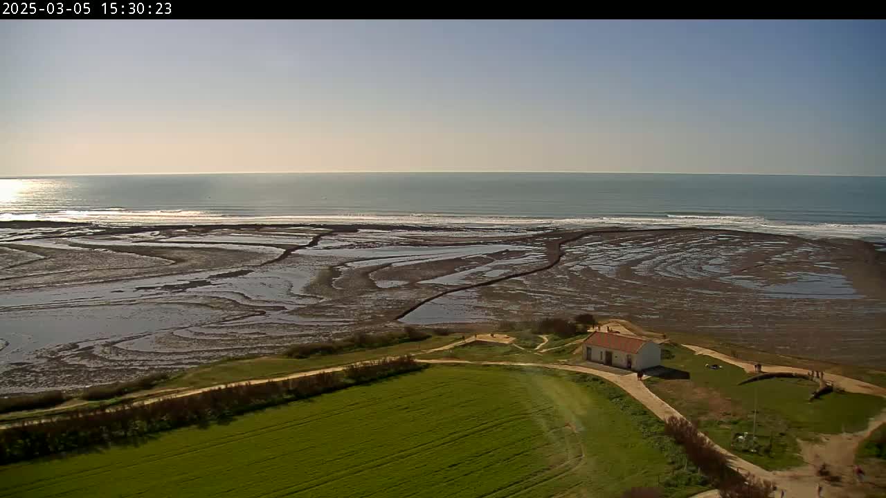 A small white building sits on a grassy hill overlooking a tidal flat and a calm ocean under a clear sky.
