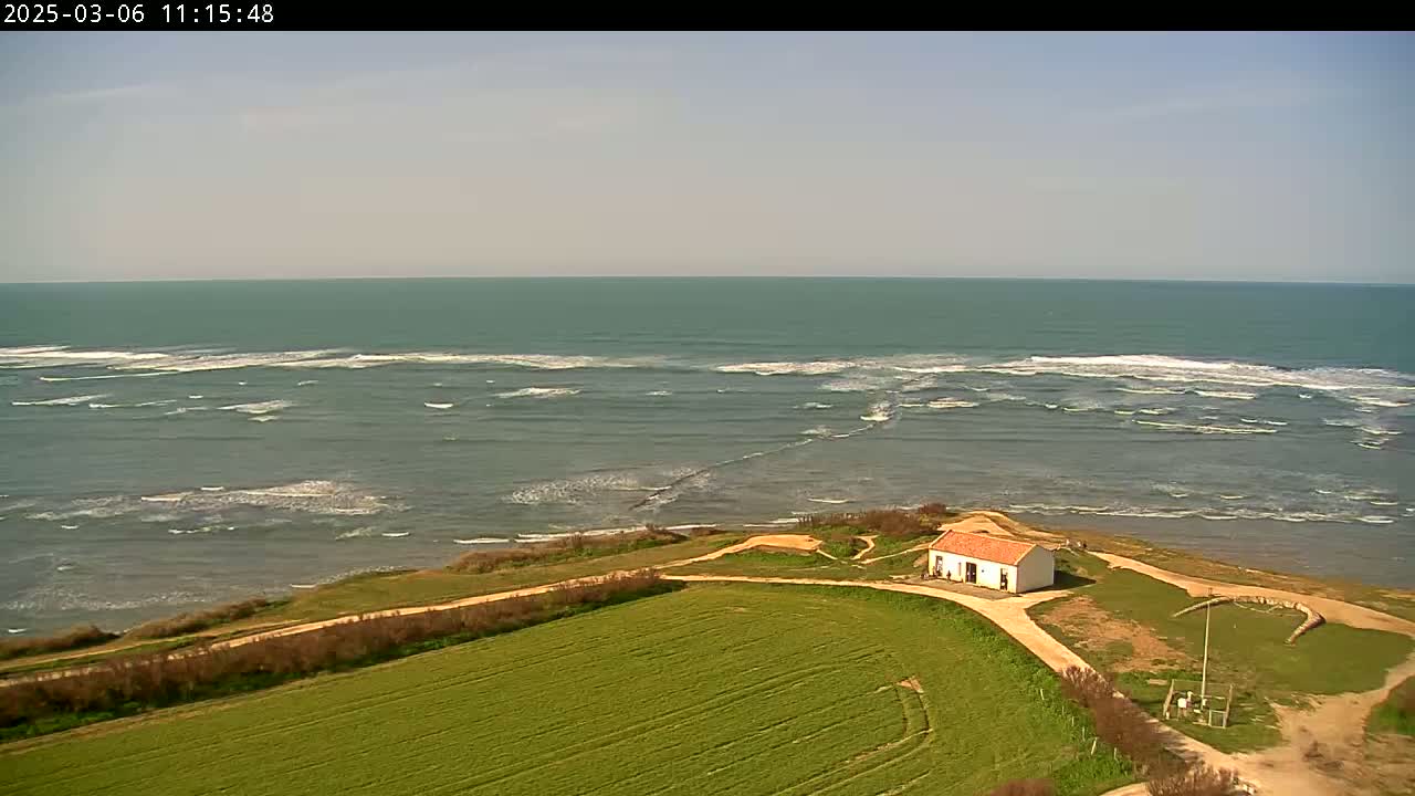 A small, white building with a red roof sits on a grassy hill overlooking a calm sea with small waves under a clear sky.