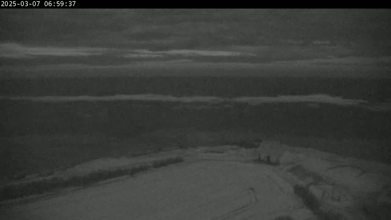 A low-light, nighttime image shows a snow-covered landscape with a few structures visible in the foreground under a cloudy sky.