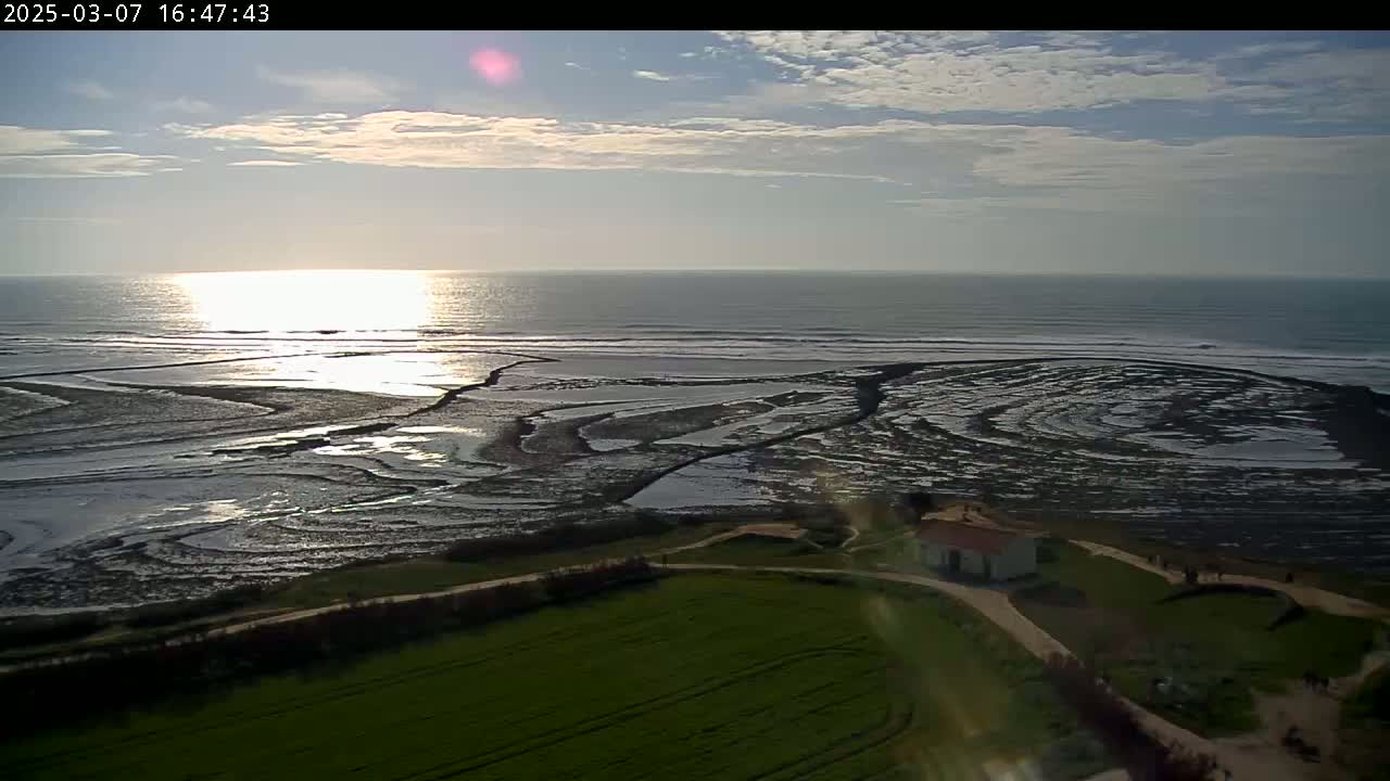 A partly cloudy sunset over a calm ocean and tidal flats, with a small building and green field visible in the foreground.