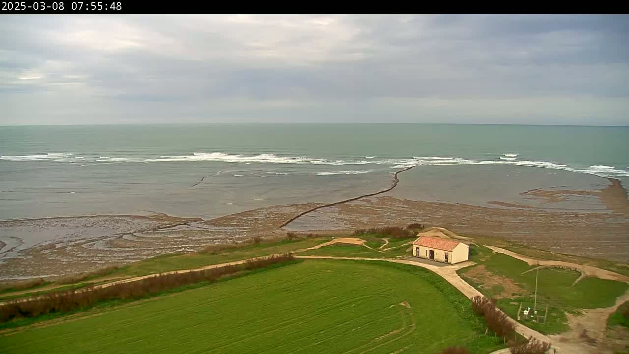 An aerial view shows a small building on a grassy hillside overlooking a calm sea with small waves under an overcast sky.