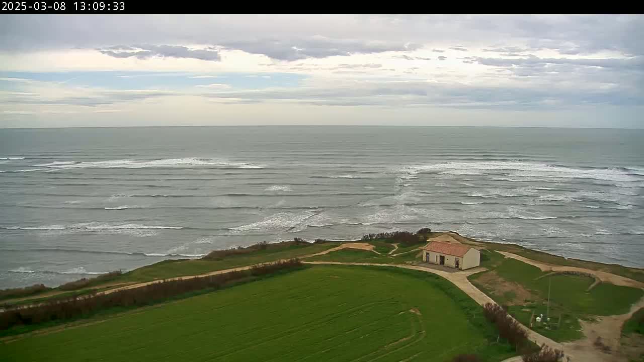 An aerial view shows a small building on a grassy hillside overlooking a grey ocean with small waves under a partly cloudy sky.