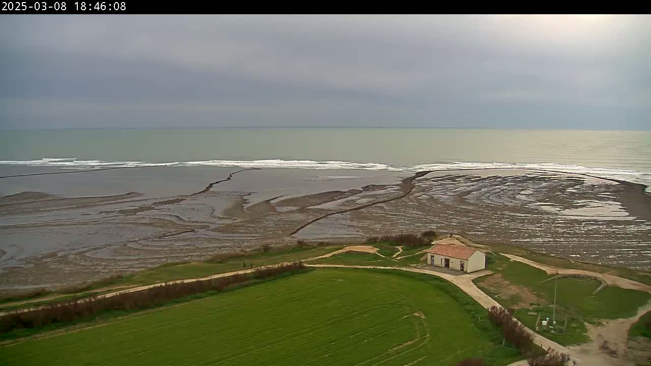 An aerial view shows a small building on a grassy area overlooking a rocky, tidal shore and a calm, overcast sea.