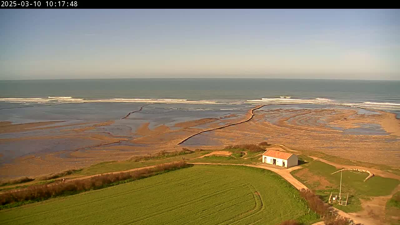 A small house sits on a grassy hill overlooking a sandy beach and ocean with gentle waves under a clear sky.