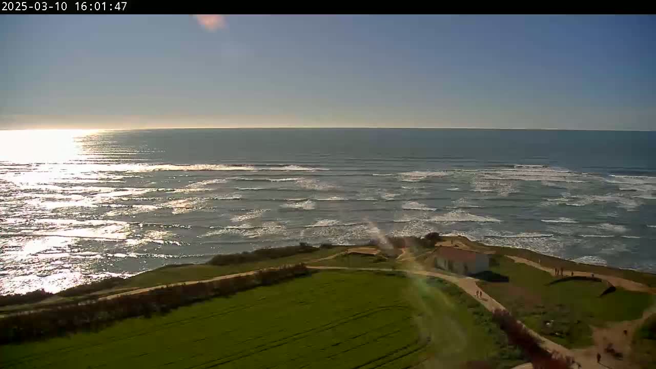 A high-angle view shows a mostly sunny day overlooking a coastline with gentle waves, a small building, and green fields.
