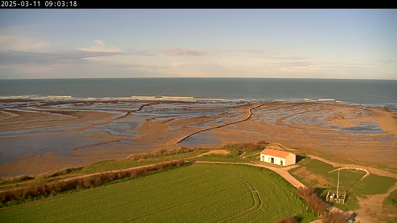 An aerial view shows a small building near a green field overlooking a beach with exposed tidal flats and a calm ocean under a mostly clear sky.