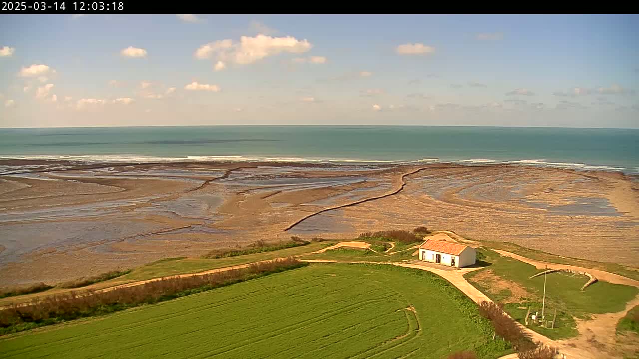 An aerial view shows a small, white building situated on a grassy hill overlooking a tidal flat and a calm ocean under a partly cloudy sky.