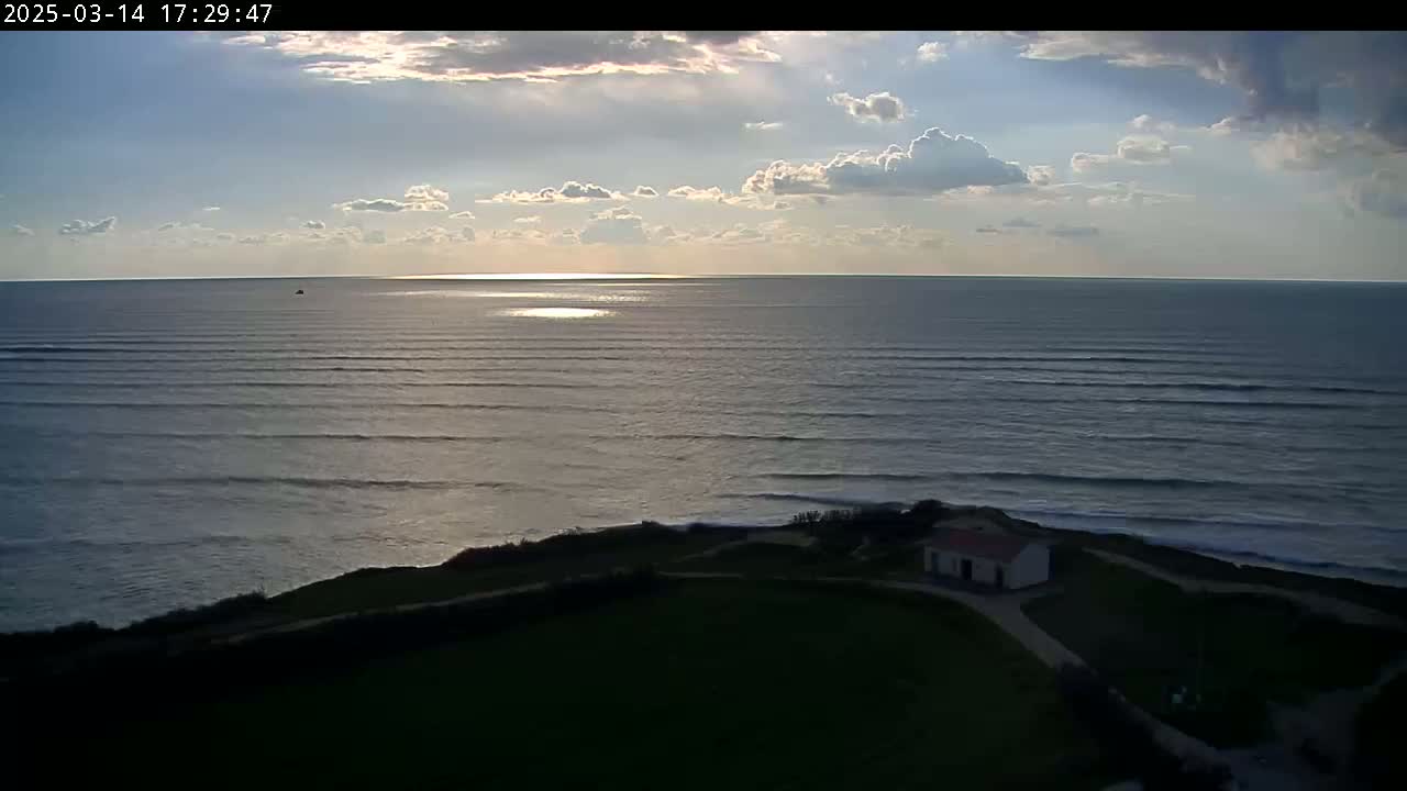 A small building sits on a grassy cliff overlooking a calm ocean under a partly cloudy sky.