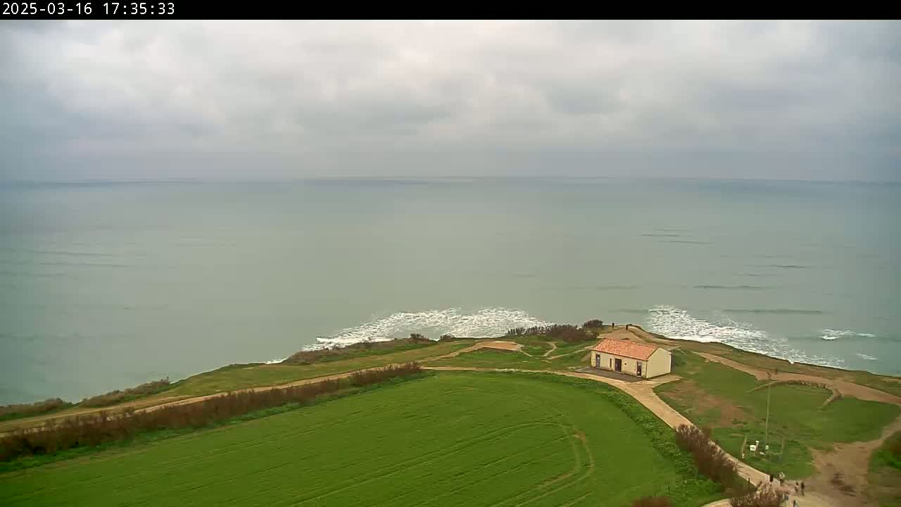 A small building sits on a grassy cliff overlooking a calm, grey sea under an overcast sky.