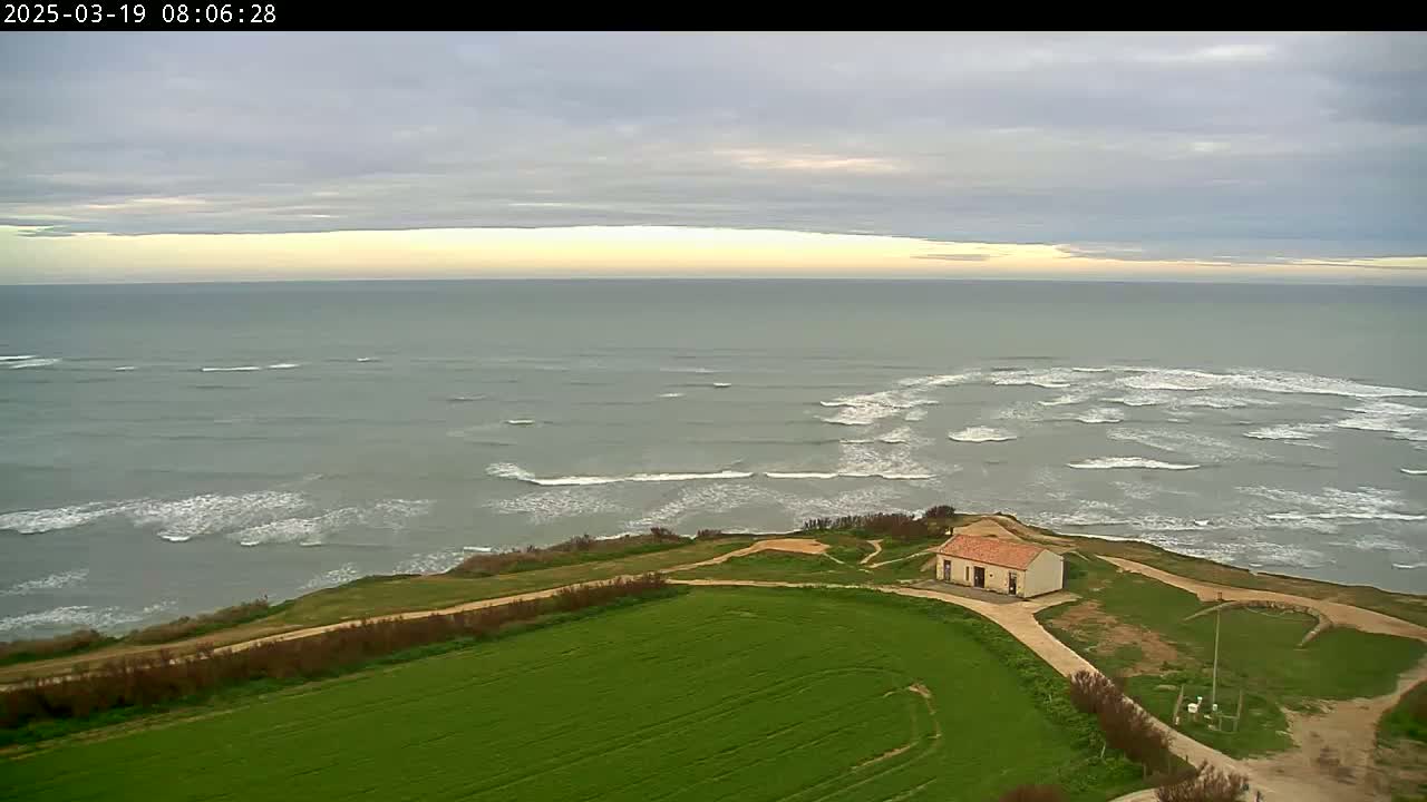A small building sits on a grassy coastal area overlooking a grey ocean with small waves under an overcast sky.