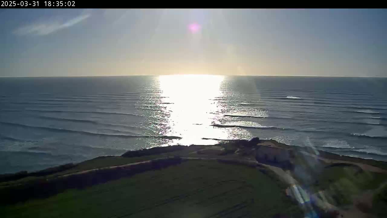 A high-angle, long shot shows a bright, sunny day overlooking a calm ocean with small waves rolling towards the shore, where a small building sits on a grassy cliff.