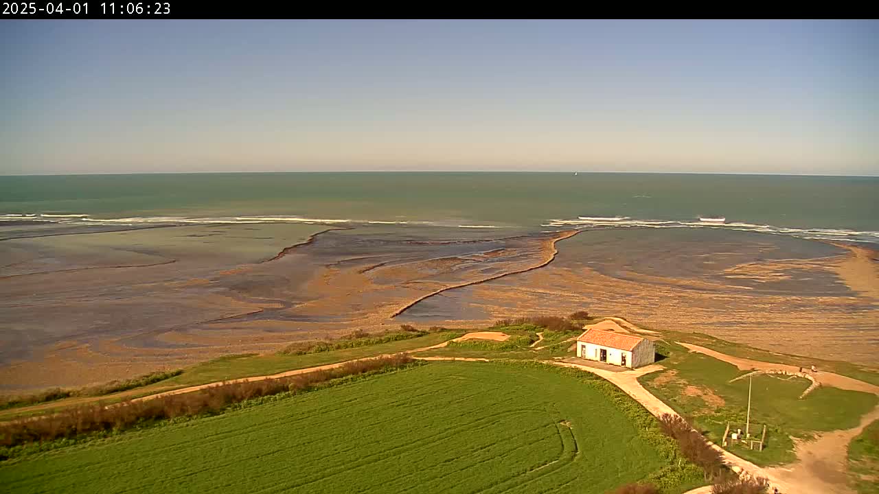 A small white building sits on a grassy hill overlooking a rocky, tidal shoreline and a calm, light-green ocean under a clear, sunny sky.