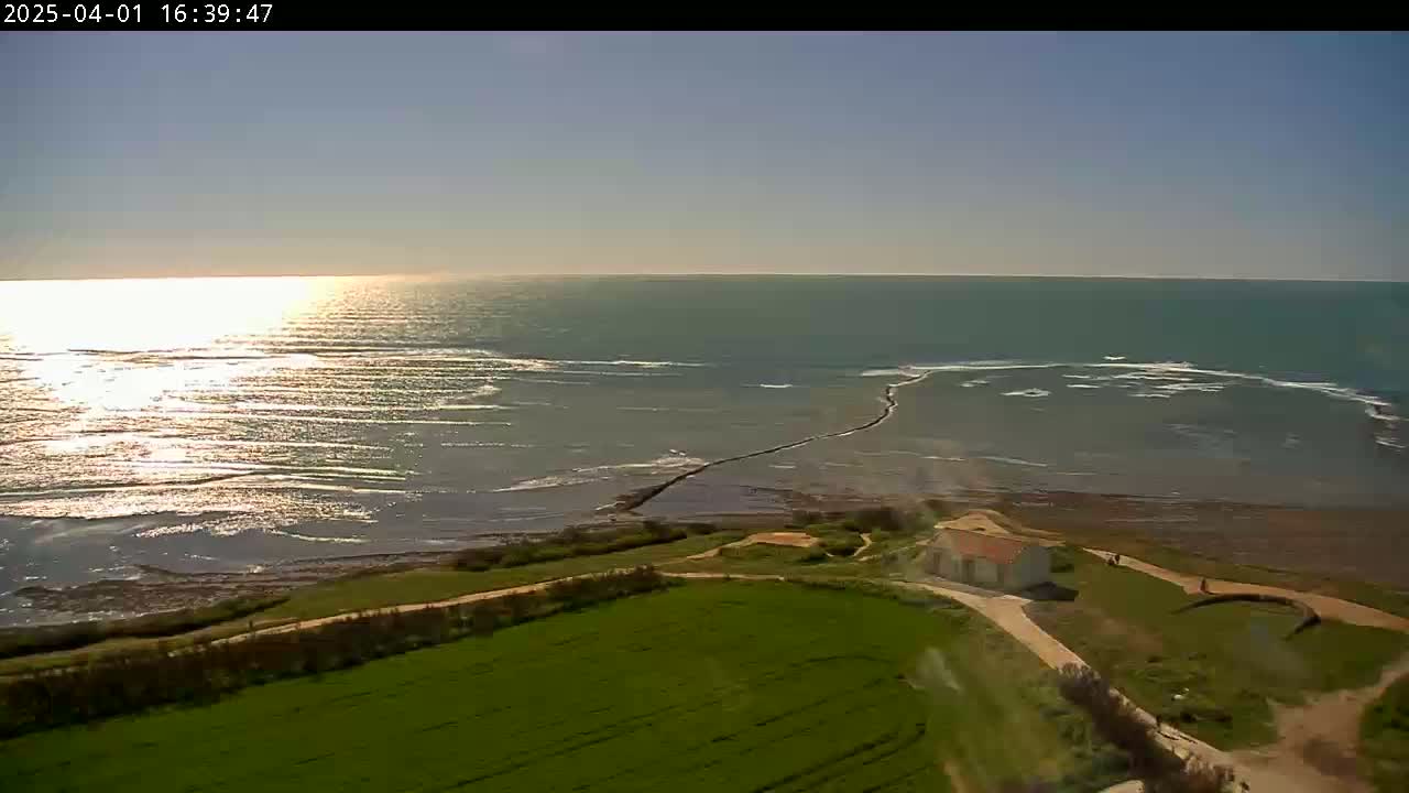 A high-angle view shows a calm, sunlit sea meeting a grassy coastline with a small building and a paved pathway, under a clear sky.