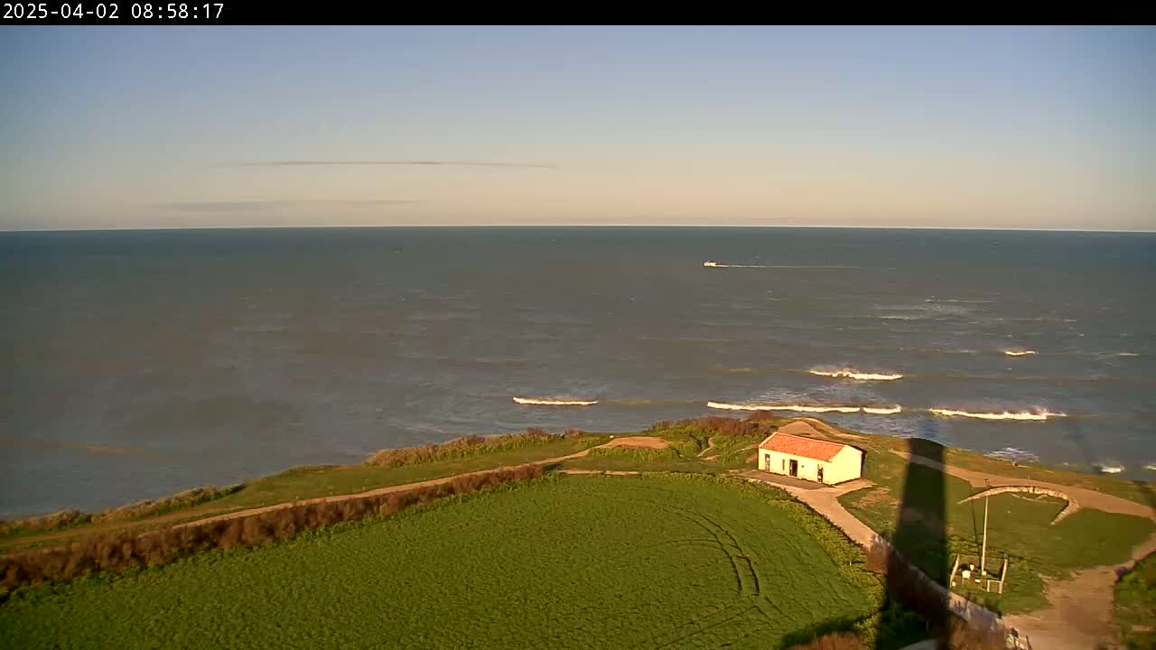 A small, red-roofed building sits on a grassy cliff overlooking a calm sea with small waves under a clear, light-blue sky.