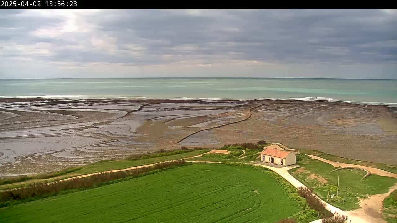 A small house sits on a grassy hill overlooking a wide expanse of exposed, textured mudflats leading to a calm, light-green ocean under a mostly cloudy sky.