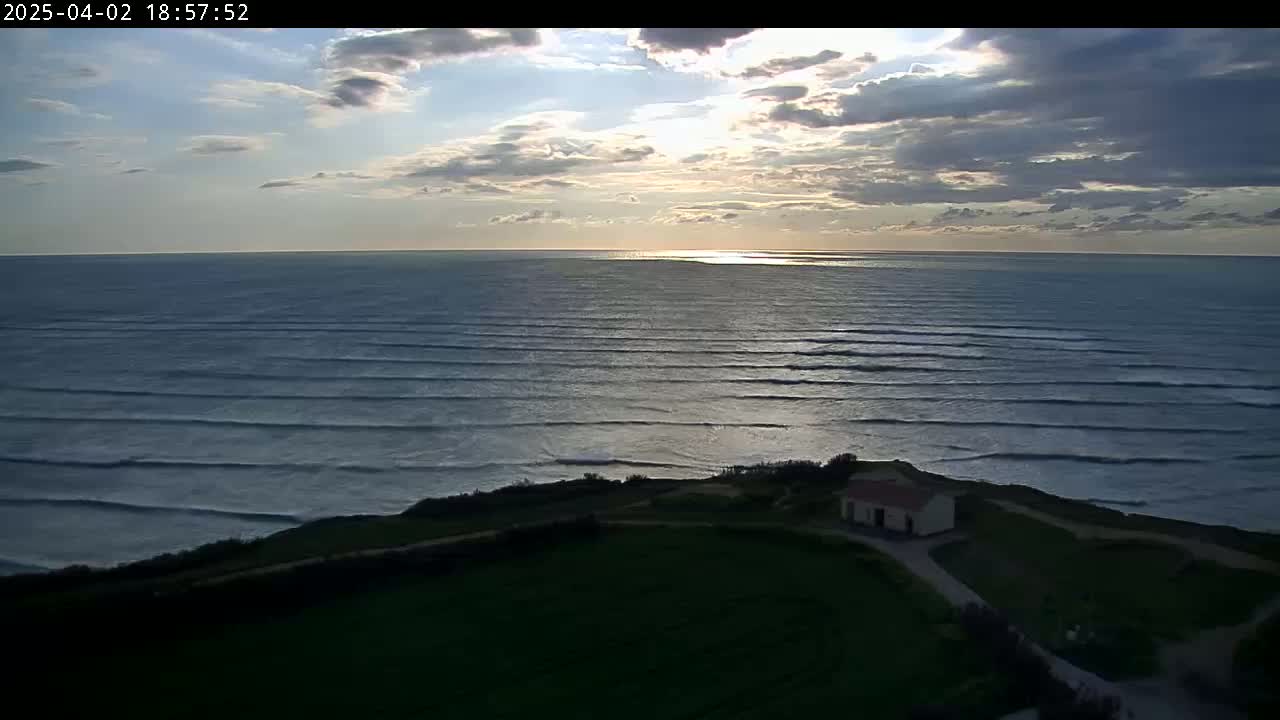 A small building sits on a grassy cliff overlooking a calm ocean with gently rolling waves under a partly cloudy sunset sky.