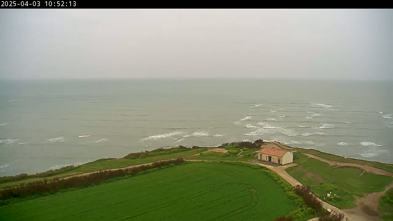A small building sits on a grassy cliff overlooking a gray, wavy ocean under an overcast sky.