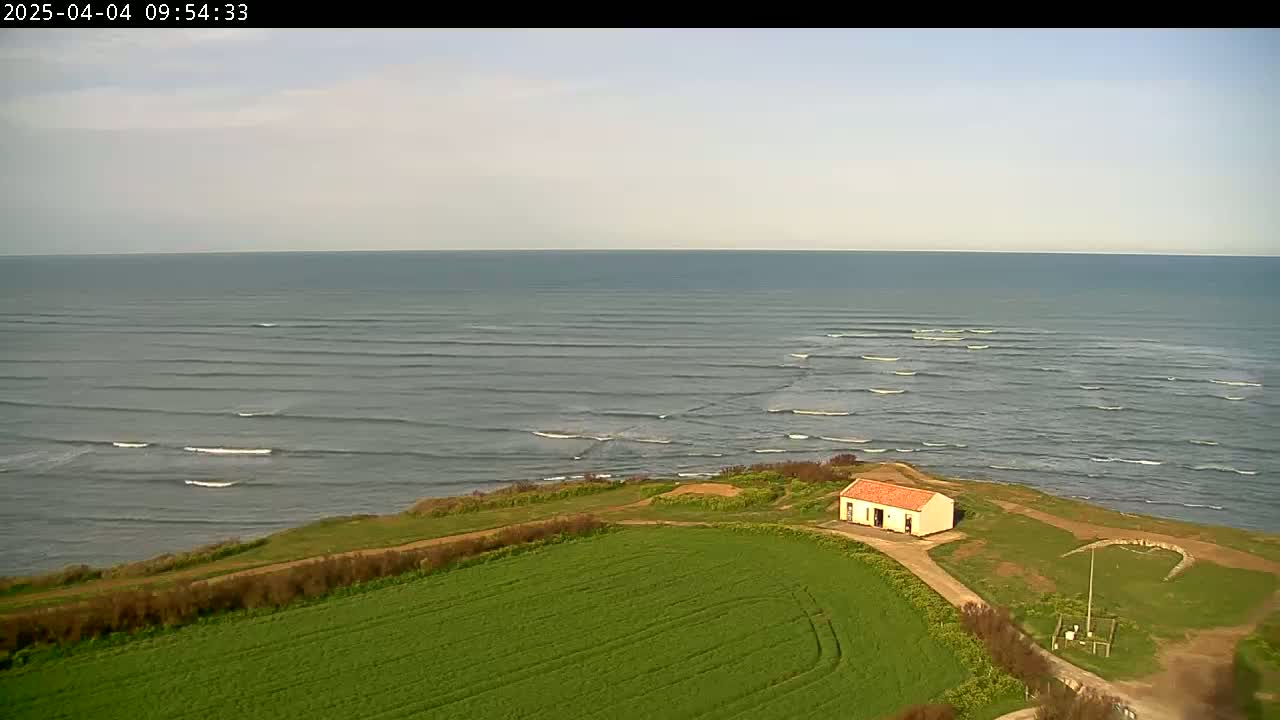 A small, red-roofed building sits on a grassy cliff overlooking a calm ocean with small, rolling waves under a mostly clear sky.